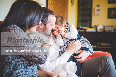 Shy female toddler sitting on fathers lap in living room