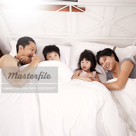 Young Chinese family of parents and two young children laying in bed together at home