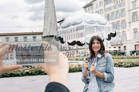 Young woman holding umbrella posing for friend using smartphone to take photograph, Piazza Santa Maria Novella, Florence, Tuscany, Italy