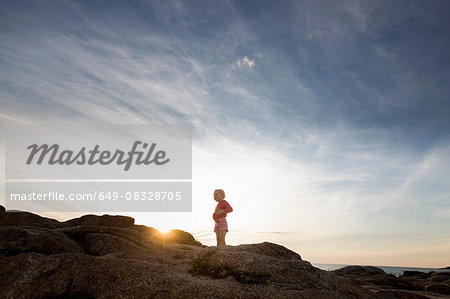 Silhouetted female toddler on coastal rock at sunset, Calvi, Corsica, France