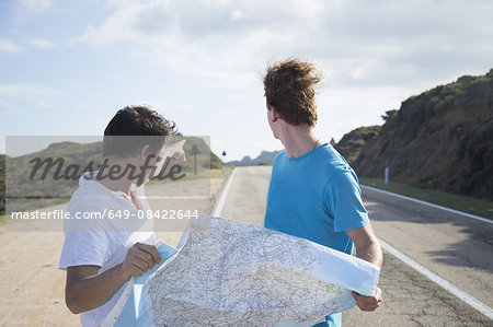 Young men on road holding map, looking over shoulder pointing, Costa Smeralda, Sardinia, Italy