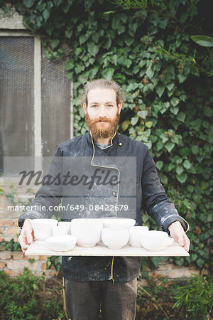 Front view of bearded mid adult man holding tray with clay pot looking at camera smiling