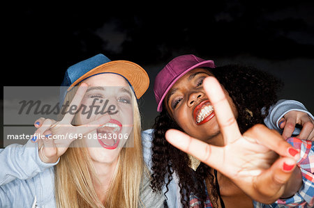 Young woman wearing baseball cap arm around friend looking at camera doing peace sign