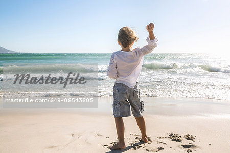 Full length rear view of boy on beach throwing stones into ocean