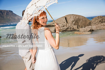 Couple holding hands, walking on coastline holding umbrella looking over shoulder at camera smiling