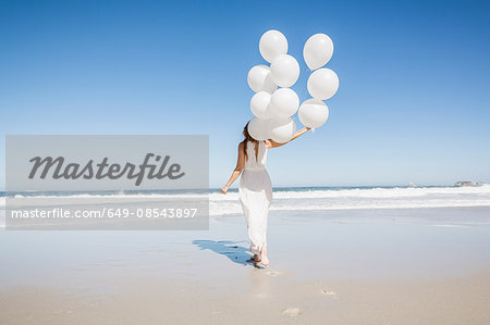 Full length rear view of woman on beach wearing white dress holding balloons