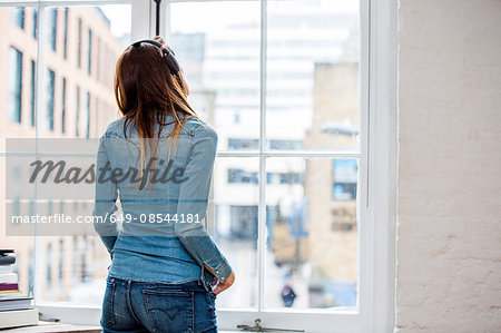 Rear view of young woman listening to music looking through city apartment window