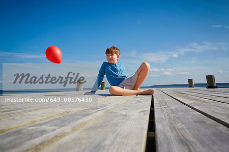 Young boy sitting on wooden pier, holding red helium balloon