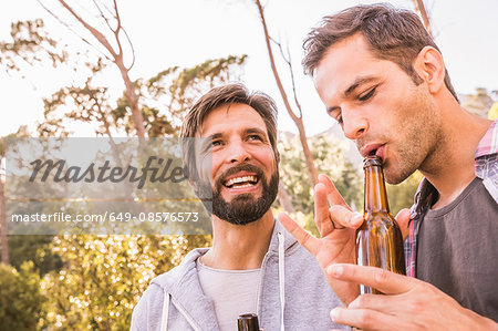 Two men blowing into beer bottle whilst camping in forest, Deer Park, Cape Town, South Africa