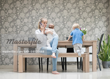 Mother using smartphone whilst holding baby boy, older son sitting beside her Mother using smartphone whilst holding baby boy, older son sitting beside her