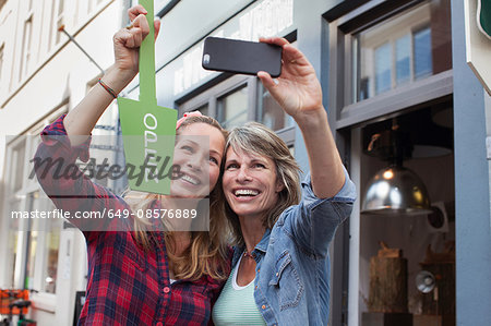 Women in front of shop holding open sign taking selfie with smartphone