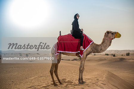 Young woman wearing traditional middle eastern clothes riding camel in desert, Dubai, United Arab Emirates