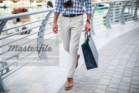 Waist down view of young man strolling on waterfront carrying shopping bag, Dubai, United Arab Emirates
