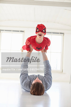 Father lying on floor holding up son wearing superhero costume