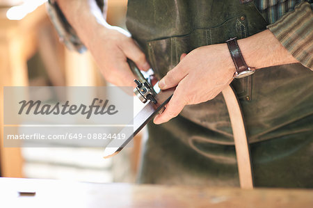 Male worker in leather workshop, punching holes in leather belt, mid section, close-up