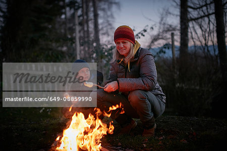 Mature woman and son toasting marshmallows on garden campfire at dusk
