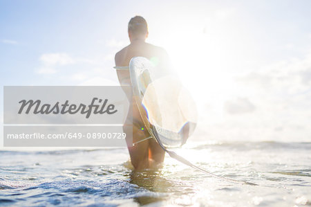 Rear view of woman carrying surfboard in sunlit sea, Nosara, Guanacaste Province, Costa Rica