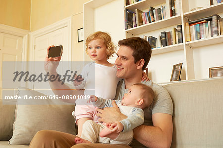 Mid adult man taking smartphone selfie with toddler and baby daughter on sofa