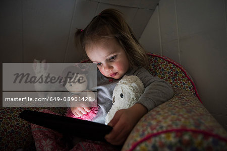 Female toddler sitting up in bed staring at digital tablet