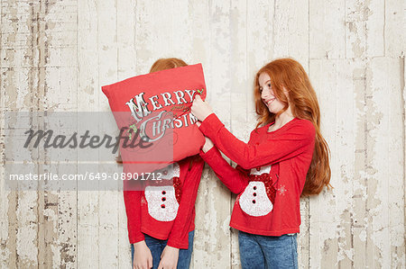 Portrait of twin sisters wearing Christmas jumpers, one sister covering sister's face with cushion