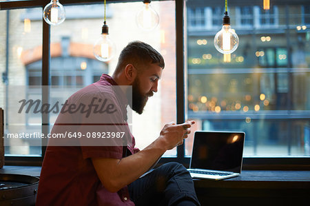 Young male hipster looking at smartphone in cafe window seat