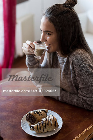 Young woman sitting at table, drinking coffee, pastry on table in front of her