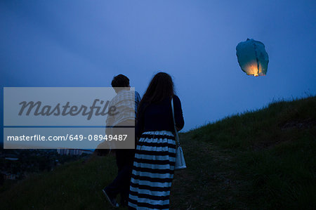 Couple walking up grassy hill at dusk, watching sky lantern in air, rear view