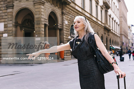 Mature woman with long grey hair with wheeled suitcase hailing a cab in Florence, Italy