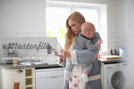 Mother standing in kitchen, holding baby boy, looking at digital tablet