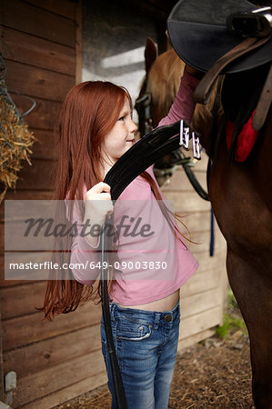 Girl fastening horse's saddle at barn