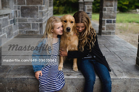 Girl and her sister sitting on patio hugging their golden retriever
