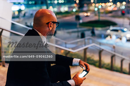 Mature businessman outdoors at night, sitting on steps, holding smartphone