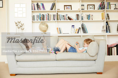 Pregnant woman reclining on sofa with smartphone opposite daughter reading storybook