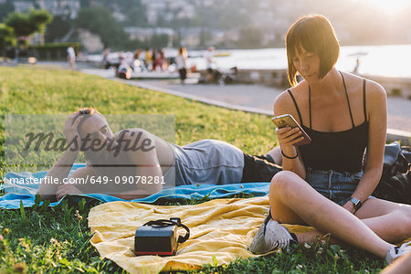 Young couple looking at smartphone on waterfront grass, Lake Como, Lombardy, Italy