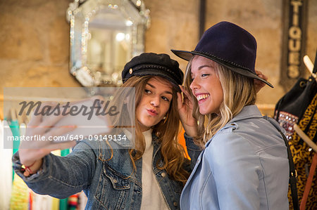 Two friends in shop, trying on hats, taking selfie, using smartphone