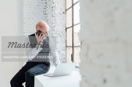 Businessman sitting on windowsill using laptop and smartphone