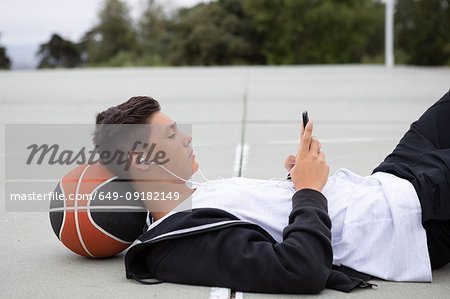 Male teenage basketball player lying on basketball court looking at smartphone
