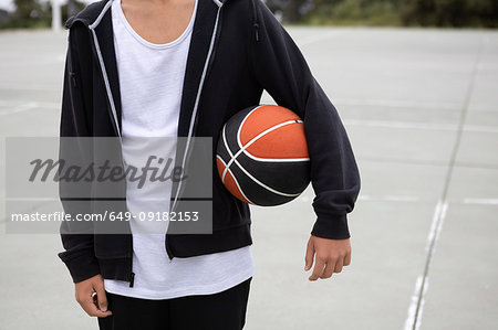 Male teenage basketball player on basketball court with ball under his arm, mid section