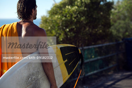 Mid adult male surfer carrying surfboard along beach walkway, rear view, Camps Bay Beach, Cape Town, South Africa