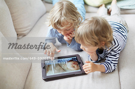 Two boys lying on fronts side by side on sofa using digital tablet