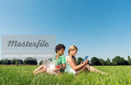 Girl and boy sitting back to back in field using digital tablets