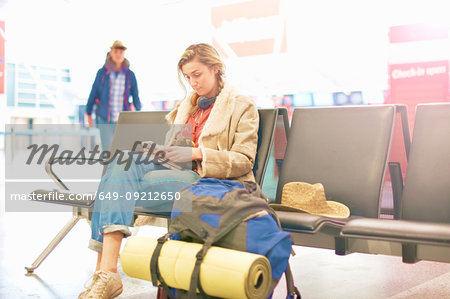 Young woman at airport, sitting with backpack beside her, using smartphone