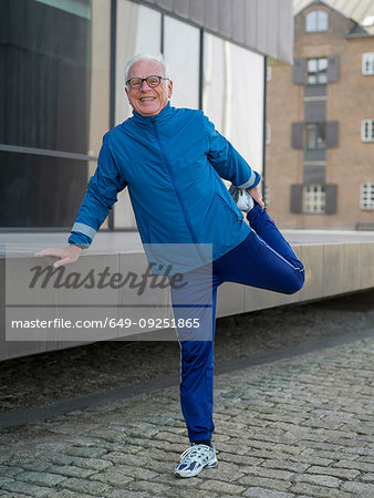 Senior man in tracksuit doing warmup exercises on cobbled street, portrait, Copenhagen, Hovedstaden, Denmark