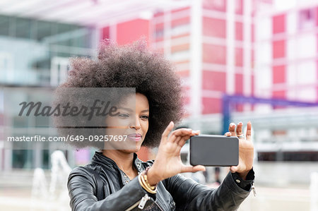 Young woman with afro hair at city train station, taking smartphone selfie