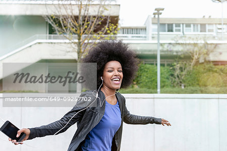 Happy young woman with afro hair in city, laughing and dancing to smartphone music