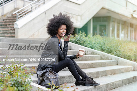 Young woman with afro hair sitting on city stairway, talking to smartphone