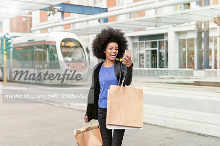 Young woman with afro hair at city train station carrying shopping bags, taking smartphone selfie