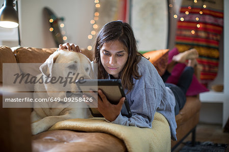 Labrador retriever and young woman lying on living room sofa looking at digital tablet