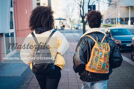 Two young women carrying backpacks strolling on urban sidewalk, rear view Two young women carrying backpacks strolling on urban sidewalk, rear view