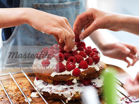 Girl and her sister baking a cake, decorating cake with fresh cream and raspberries at kitchen table, cropped view of hands